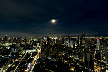 Obraz premium A breathtaking night view of Bangkok's skyline illuminated by city lights and a full moon. The cityscape showcases the blend of modern skyscrapers and vibrant urban life, reflecting the city's dynamic