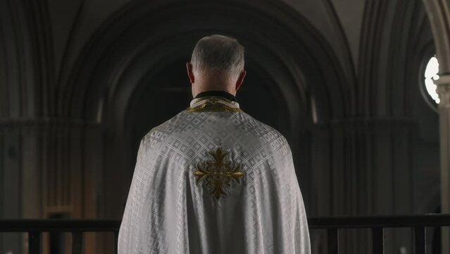 Back view zoom off shot of pastor in white chasuble standing on balcony inside of cathedral