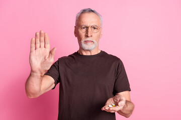 Portrait of serious senior man with beard dressed brown t-shirt in glasses give you pills show palm isolated on pink color background