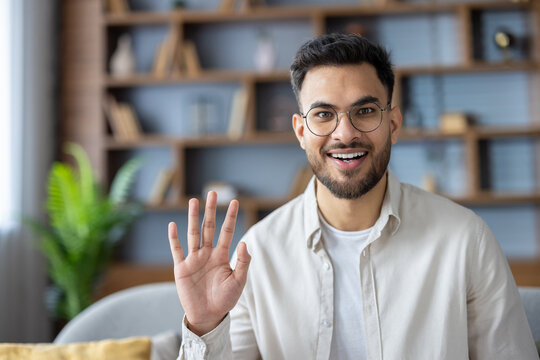 Close-up portrait of a young Indian man sitting on the sofa at home wearing glasses and casual clothes, greeting, smiling and waving at the camera - Powered by Adobe