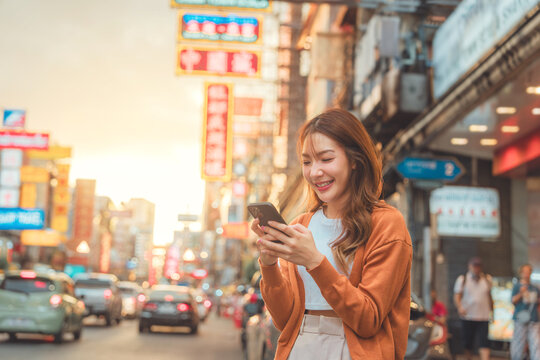 Happy young Asian tourist woman using smartphone on street, Female traveller in Yaowarat Chinatown street food market during sunset in Bangkok, Thailand. - Powered by Adobe