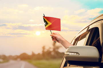 Waving the East Timor flag against the sunrise or sunset from a car driving along a country road. Holding the East Timor flag, traveling by car, on a weekend trip.