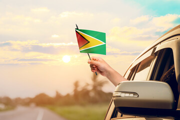 Waving the Guyana flag against the sunrise or sunset from a car driving along a country road. Holding the Guyana flag, traveling by car, on a weekend trip.