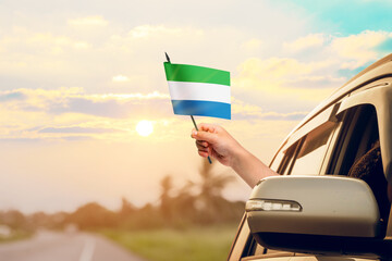 Waving the Sierra Leone flag against the sunrise or sunset from a car driving along a country road. Holding the Sierra Leone flag, traveling by car, on a weekend trip.