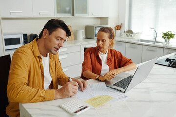 Man calculating the bills while his wife using laptop, they sitting in the kitchen and discussing...