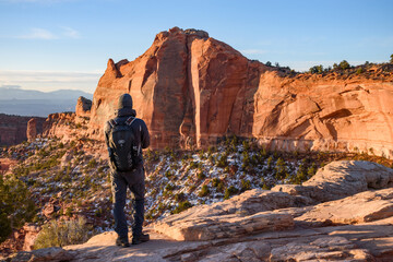 A male hiker stands at the edge of a cliff in Utah's Canyonlands National Park, admiring the breathtaking view of the snow-dusted landscape and rugged red rock formations at sunrise - USA