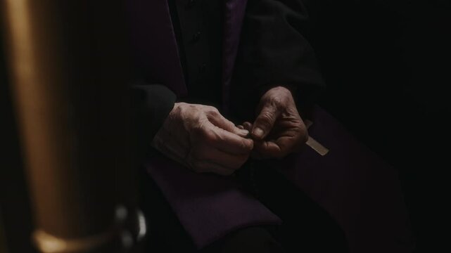 Close-up view of hands of male priest in cassock sitting in dark confession booth and holding rosary beads