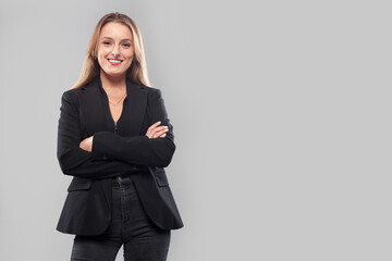 Happy female teacher in formal suit standing on gray background at studio while looking at camera with arms crossed.