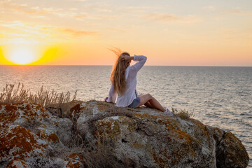 A woman with long hair sits on the rocks on the seashore at sunset. Travel and tourism