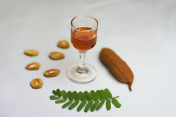 Taça de vidro pequena com licor dentro fundo branco acinzentado desfocado rodeado por uma fruta de tamarindo madura marrom e alguns gomos da fruta cortados em postas finas e uma folha verde 