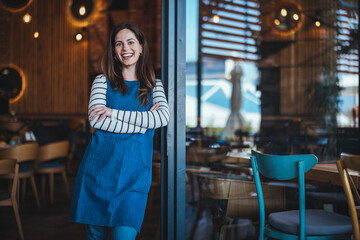 A cheerful female bartender in an apron stands with crossed arms in a cozy coffee house, surrounded by wooden interior elements.