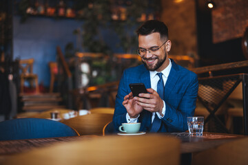 A cheerful man in a tailored suit engages with his phone at a cafe, a coffee cup and glass of water on his table, embodying modern connectivity and business on-the-go.