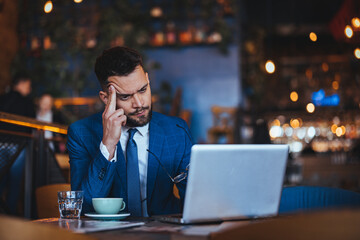 A pensive Caucasian man in a suit engages with a laptop in a bustling cafe, displaying signs of worry while sipping coffee beside a glass of water.