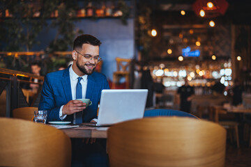 Professional Caucasian male in formal attire works on a laptop while enjoying a cup of coffee inside a cozy coffee shop.