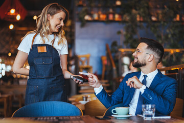 Smiling man in a suit engages with a waitress, using a card for payment, reflecting a casual yet professional dining transaction in a cafe setting.