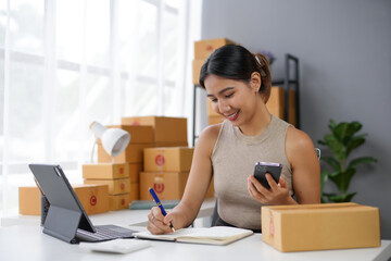 Smiling businesswoman managing online orders in a modern office with packaging boxes, laptop, and mobile phone. E-commerce concept.