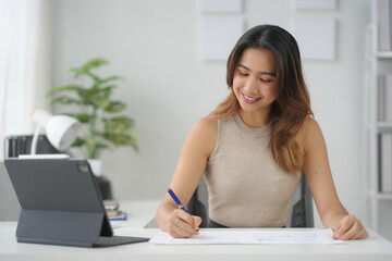 Smiling woman working from home, writing in notebook, using tablet computer, in modern light office with greenery.