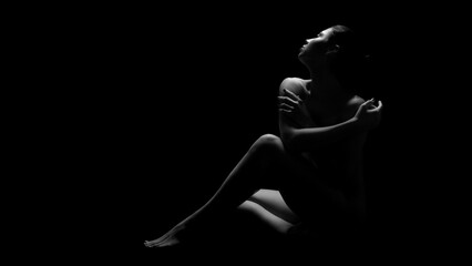 A black and white photograph of a woman sitting in darkness, illuminated only by a single light source. She has her arms crossed, and her gaze is directed upwards.