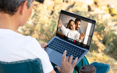 A senior woman sits outdoors, using a laptop to video chat with a young couple. The woman is...