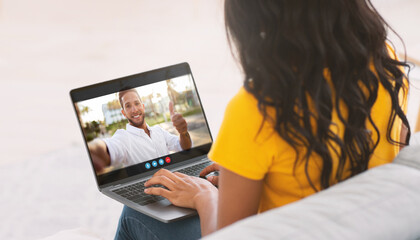Black woman is seated on a couch, video chatting with a man on her laptop. The man is giving a thumbs up in the video call. She is looking at the screen and typing on the laptop keyboard.