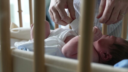 Mother’s hands carefully adjusting newborn baby’s clothing in a bassinet, viewed through crib...