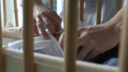 Mother’s hands carefully adjusting newborn baby’s clothing in a bassinet, viewed through crib...