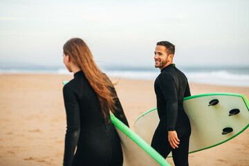 A young couple in wetsuits walk along a sandy beach with their surfboards, heading towards the water. The man is smiling and looking back at the camera, while the woman looks forward.