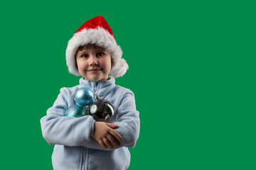 Child in Santa Claus hat with Christmas tree toys (glass balls) to decorate Christmas tree in hands on green background. Copy space.