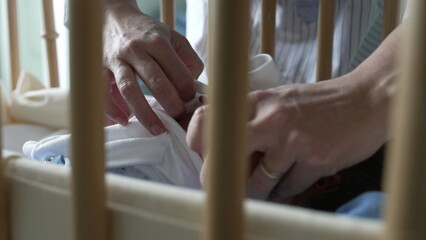 Mother’s hands tenderly dressing a newborn baby in a bassinet, visible through the crib bars,...