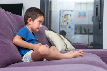 A young boy in a blue t-shirt and grey shorts is deeply engrossed in a video game while sitting on...