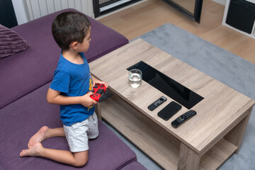 Young Boy Engaged in Video Game Kneeling on Purple Couch.
A young boy, wearing a blue t-shirt and grey shorts, is seen kneeling on a purple couch while holding a red game controller.