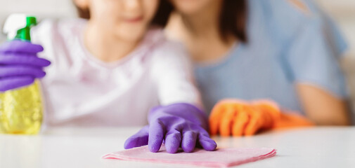 A young girl wearing purple gloves cleans a table with a pink cloth while her mother, wearing...