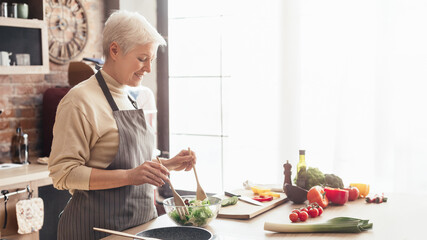A woman wearing an apron stands in a modern kitchen, preparing a fresh salad. The kitchen has a brick backsplash, white cabinets, and a large window that lets in natural light, copy space