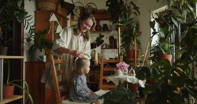 Young loving mother tying a hair bow for her adorable little daughter in grey dress sitting on a wooden chair among decorative plants in the home garden. Wide slow motion shot.