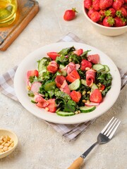 A healthy salad with cabbage kale, fresh cucumbers, radish, strawberries and smoked meat on a beige ceramic plate on a light concrete background.