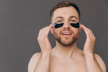 Close-up portrait of handsome bearded man applying black patches under eyes. Attractive guy caring about his appearance on grey background