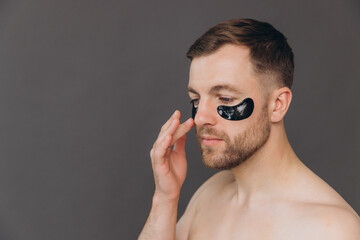 Close-up portrait of handsome bearded man applying black patches under eyes. Attractive guy caring about his appearance on grey background