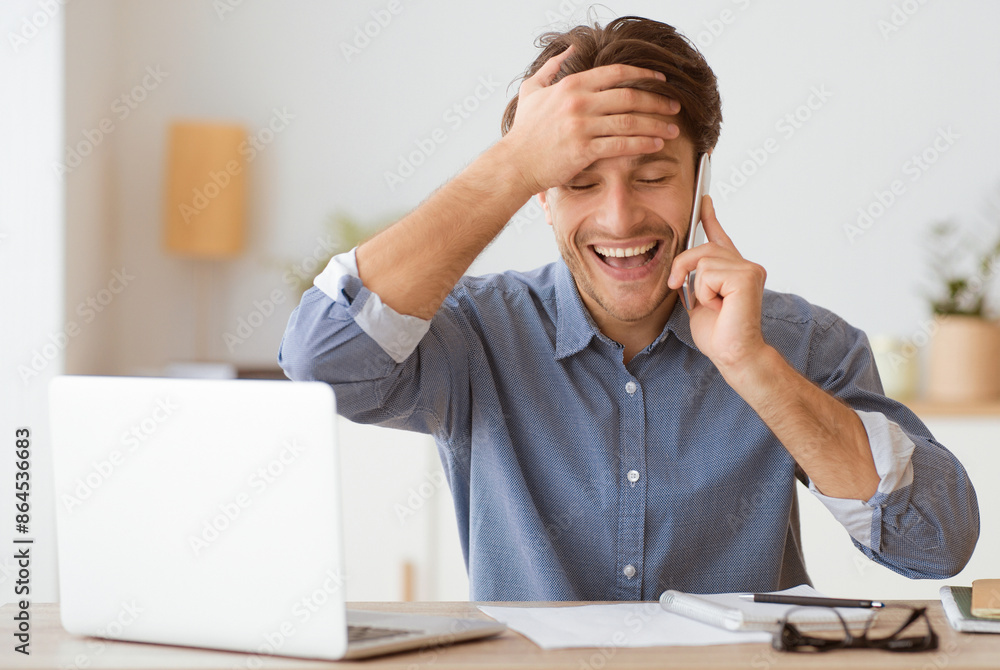 Poster a man sits at a desk with a laptop, laughing while on a phone call. he is covering his forehead with
