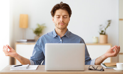 A man sits at his desk with his eyes closed, meditating. He has a laptop in front of him.