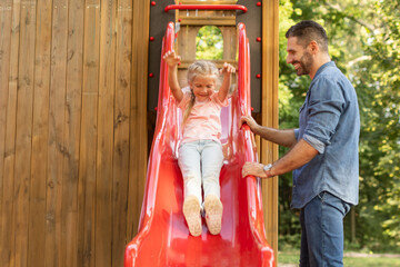 A father smiles and watches as his daughter slides down a red slide at a playground. The girl is wearing a pink shirt and jeans, and the father is wearing a blue shirt and jeans.