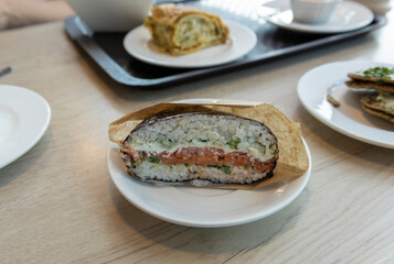 A close-up shot of a salmon and rice sandwich served on a white plate. The sandwich is wrapped in brown paper, revealing a delicious filling of salmon, rice, and other ingredients.