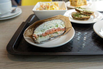 A close-up of a salmon and rice hand roll, wrapped in paper, on a white plate, sitting on a black tray in a cafe.
