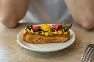 A close-up image of a pastry with yellow cream, chocolate sprinkles, and strawberries, served on a white plate in a cafe. The pastry is on a wooden table, and a fork is visible in the background.