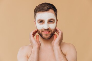 Beauty portrait of shirtless unshaved smiling man applies facial care mask against beige studio...