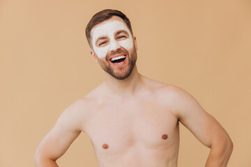 Beauty portrait of shirtless unshaved smiling man applies facial care mask against beige studio...