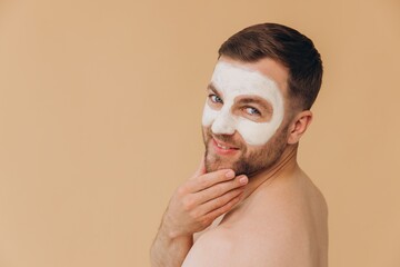 Beauty portrait of shirtless unshaved smiling man applies facial care mask against beige studio...