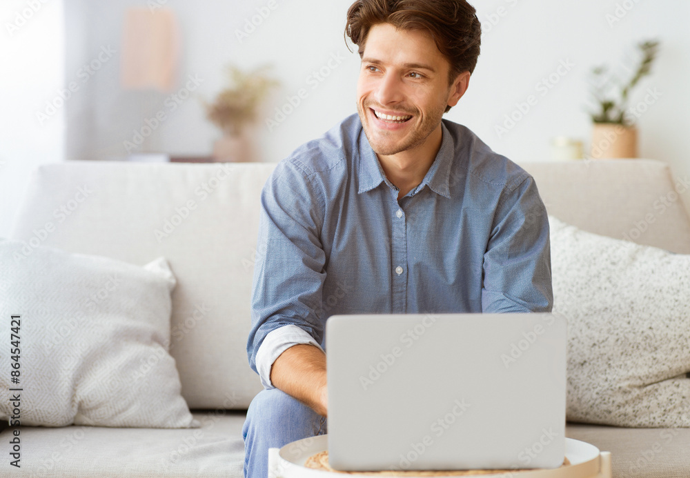 Poster A man sits on a couch in a living room, smiling while looking to the side. He is using a laptop and appears to be enjoying himself.