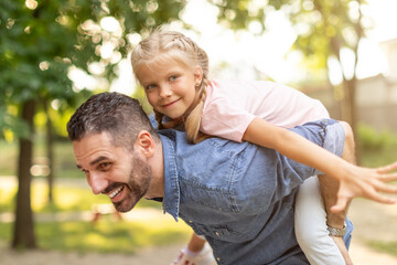 A happy father gives his daughter a piggyback ride in a summer park. The daughter is smiling and has her arms out, as if she is flying. The father is also smiling and looks very happy.