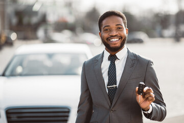 African American man in a suit stands in front of a white car, holding the keys in his right hand...