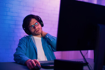 A young man in a blue shirt sits at a computer in a room with purple and blue lighting. He wears glasses and headphones and has his hand on his neck as if he is in pain. © Prostock-studio